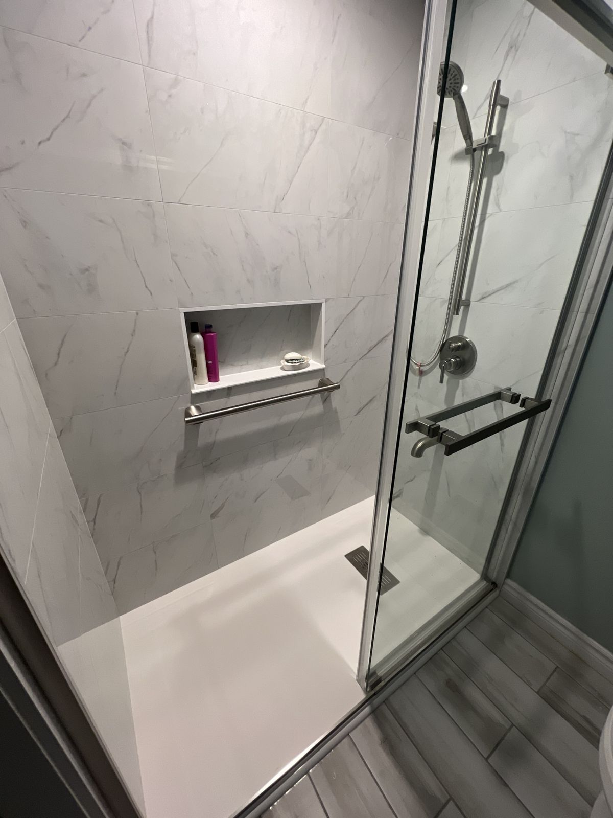 Close-up of a walk-in shower with marble-look walls, recessed niche, grab bar, slide-bar hand shower and linear drain