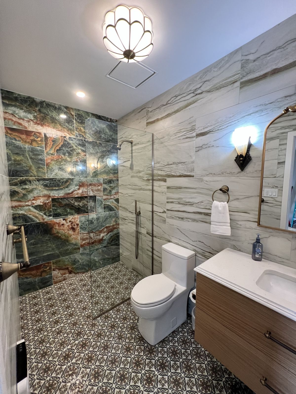 Renovated bathroom with a curbless walk-in shower in green and rust stone-look tile, patterned porcelain floor, frameless glass panel and floating wood vanity