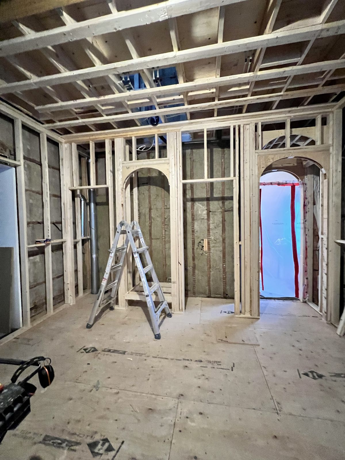 Interior framing in progress: new stud walls with two custom arched openings, insulated exterior walls and an aluminum ladder on a plywood subfloor
